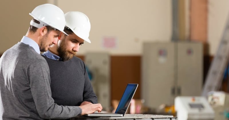 Two men wearing construction hats, looking at a laptop