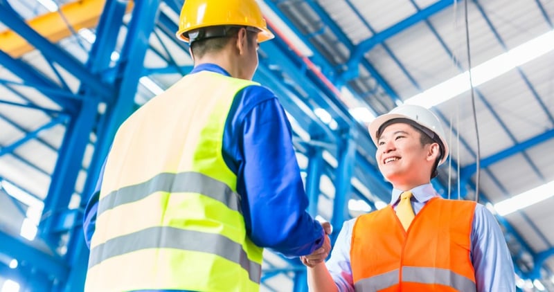 Two men wearing construction hats and high visibility vest, shaking hands in a factory